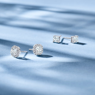 Two pairs of Royal Asscher diamond stud earrings—one round pavé and one princess cut—displayed on soft blue fabric with natural light.