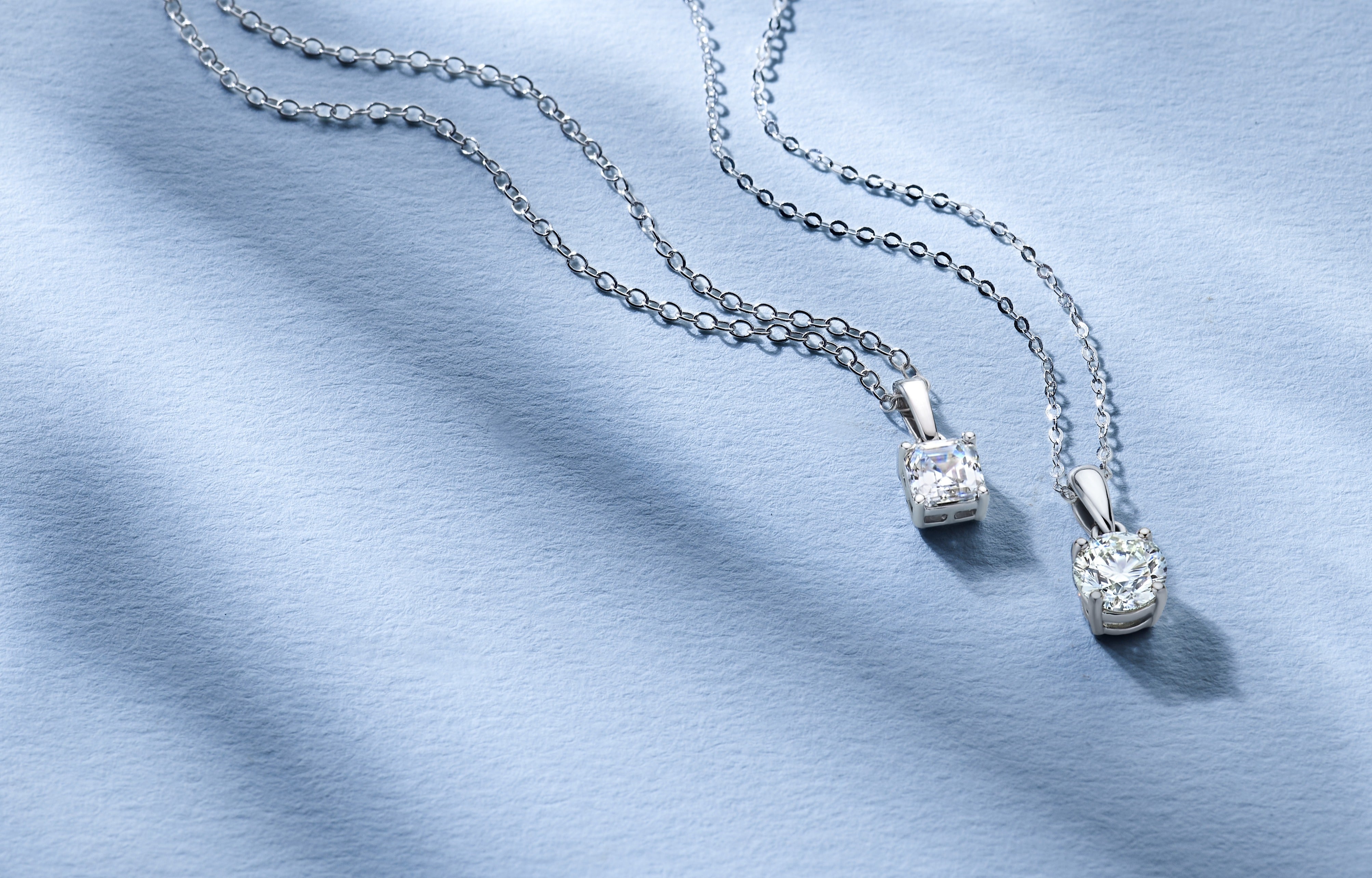 Two Royal Asscher diamond pendants with cushion-cut designs on silver chains, displayed on a soft blue fabric background.