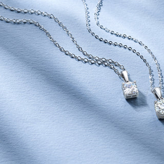 Two Royal Asscher diamond pendants with cushion-cut designs on silver chains, displayed on a soft blue fabric background.