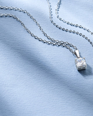Two Royal Asscher diamond pendants with cushion-cut designs on silver chains, displayed on a soft blue fabric background.