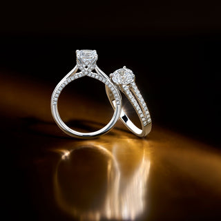 Two Royal Asscher brilliant cut diamond engagement rings with pavé bands, displayed on a reflective golden surface under soft lighting.