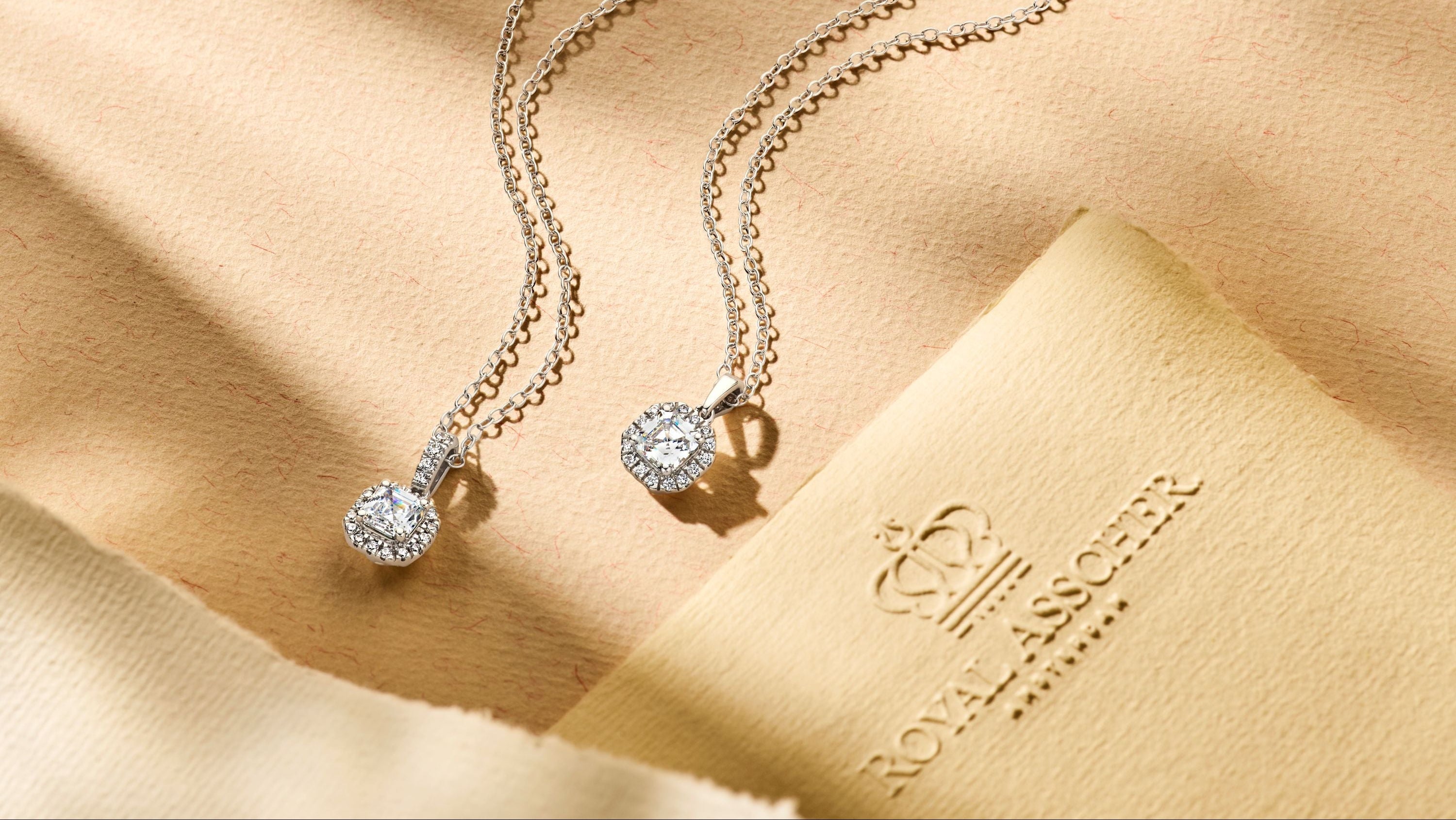 Two Royal Asscher diamond pendants with cushion-cut designs on silver chains, displayed on a soft blue fabric background.