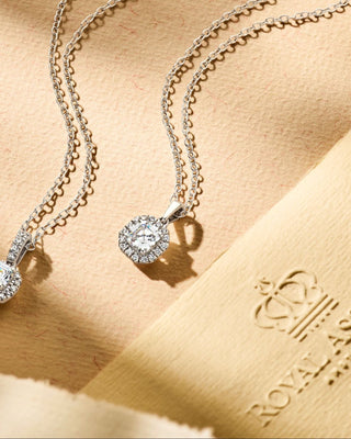 Two Royal Asscher diamond pendants with cushion-cut designs on silver chains, displayed on a soft blue fabric background.